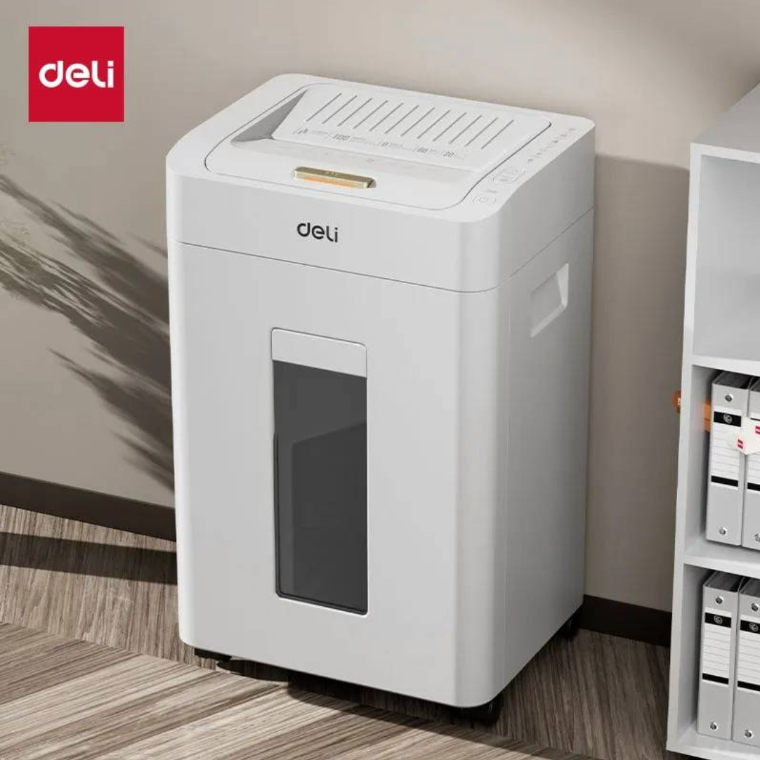 A white Deli desktop document shredder with a transparent window on the front, shown in a home office setting next to a shelf with binders.
